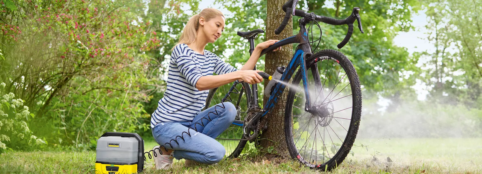 Μια γυναίκα καθαρίζει το ποδήλατό της δίπλα σε ένα δέντρο με ένα κινητό καθαριστικό εξωτερικών χώρων Kärcher