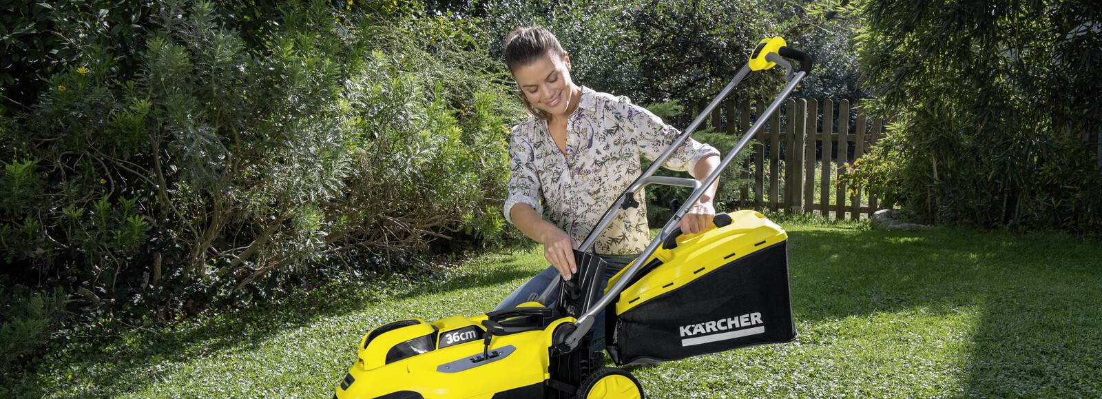 Młoda kobieta montuje kosz w akumulatorowej kosiarce do trawy. W tle bujne rośliny i fragment płotu.