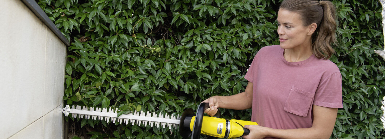 Młoda kobieta trzyma oburącz akumulatorowe nożyce do żywopłotu, którymi przycina rośliny.