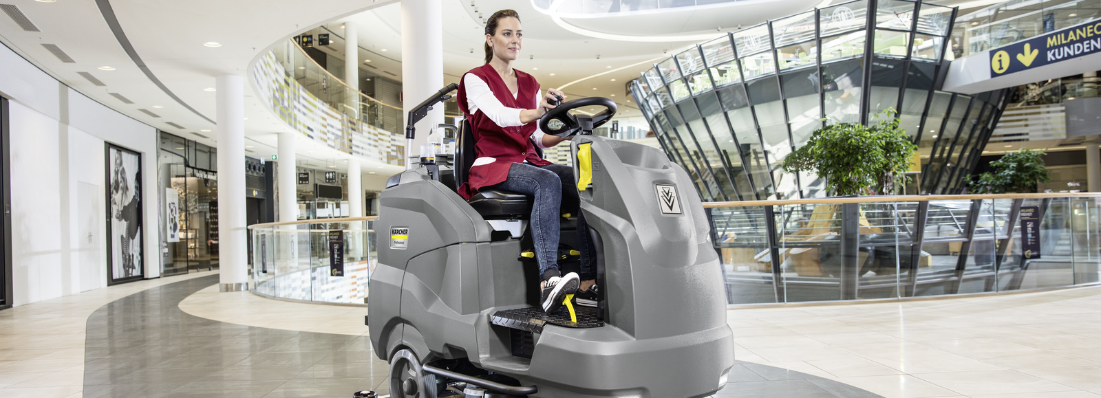 Ride-on sweeper A person cleaning the floor of a shopping center using a ride-on sweeper