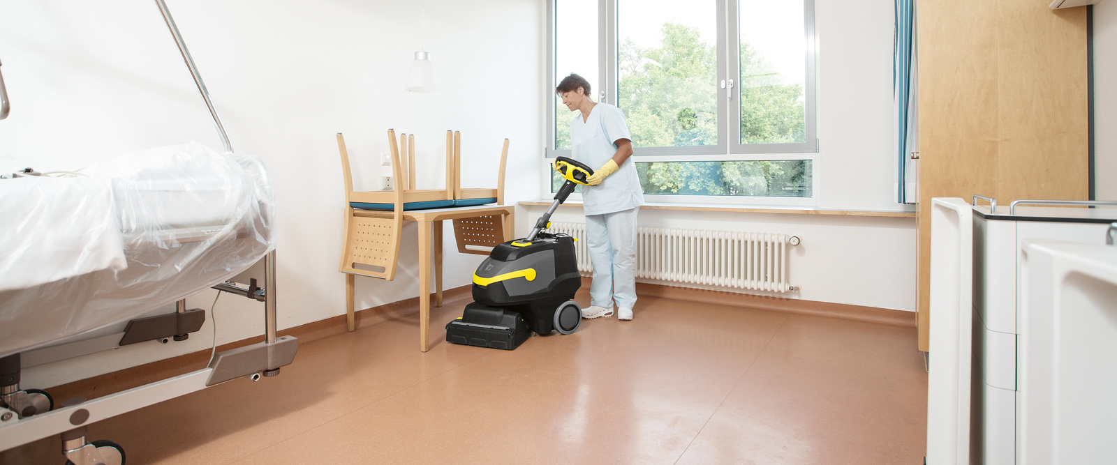 Reinigung mit der Scheuersaugmaschine Eine Person reinigt den Boden eines Patientenzimmers mit Hilfe einer Scheuersaugmaschine von Kärcher