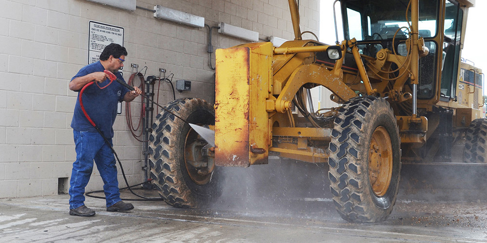 A worker pressure washes construction equipment using a Hotsy pressure washer