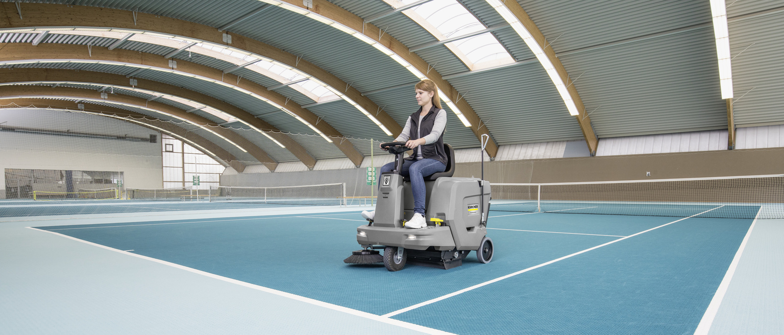 Carpet vacuum sweeper A woman cleaning the floor of an indoor tennis court with a ride-on carpet vacuum sweeper