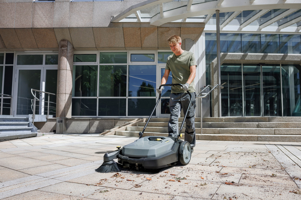 Walk-Behind Floor Sweepers A worker uses a KM 70/20 Walk-Behind Floor Sweeper outside of an office building entrance