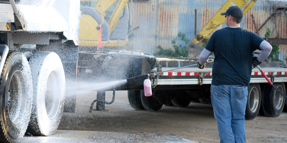 A worker sprays pressure washer detergent onto truck tires.