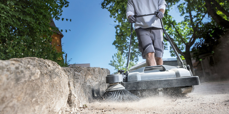 A worker pushes a walk-behind floor sweeper to clean up dirt and debris.