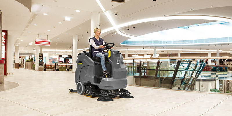 A worker cleans large floor areas using a ride-on floor scrubber