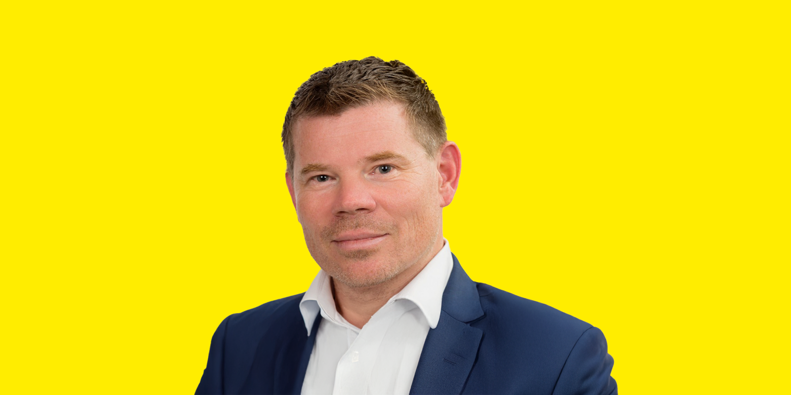 Christian Bock Das Bild zeigt ein Porträt von Christian Bock, der direkt in die Kamera blickt und leicht lächelt. Er hat kurzes, dunkelbraunes Haar und helle Augen. Er trägt ein weißes Hemd mit offenem Kragen und einen dunkelblauen Anzug. Der Hintergrund ist nur gelb, wodurch der Fokus vollständig auf der Person liegt.