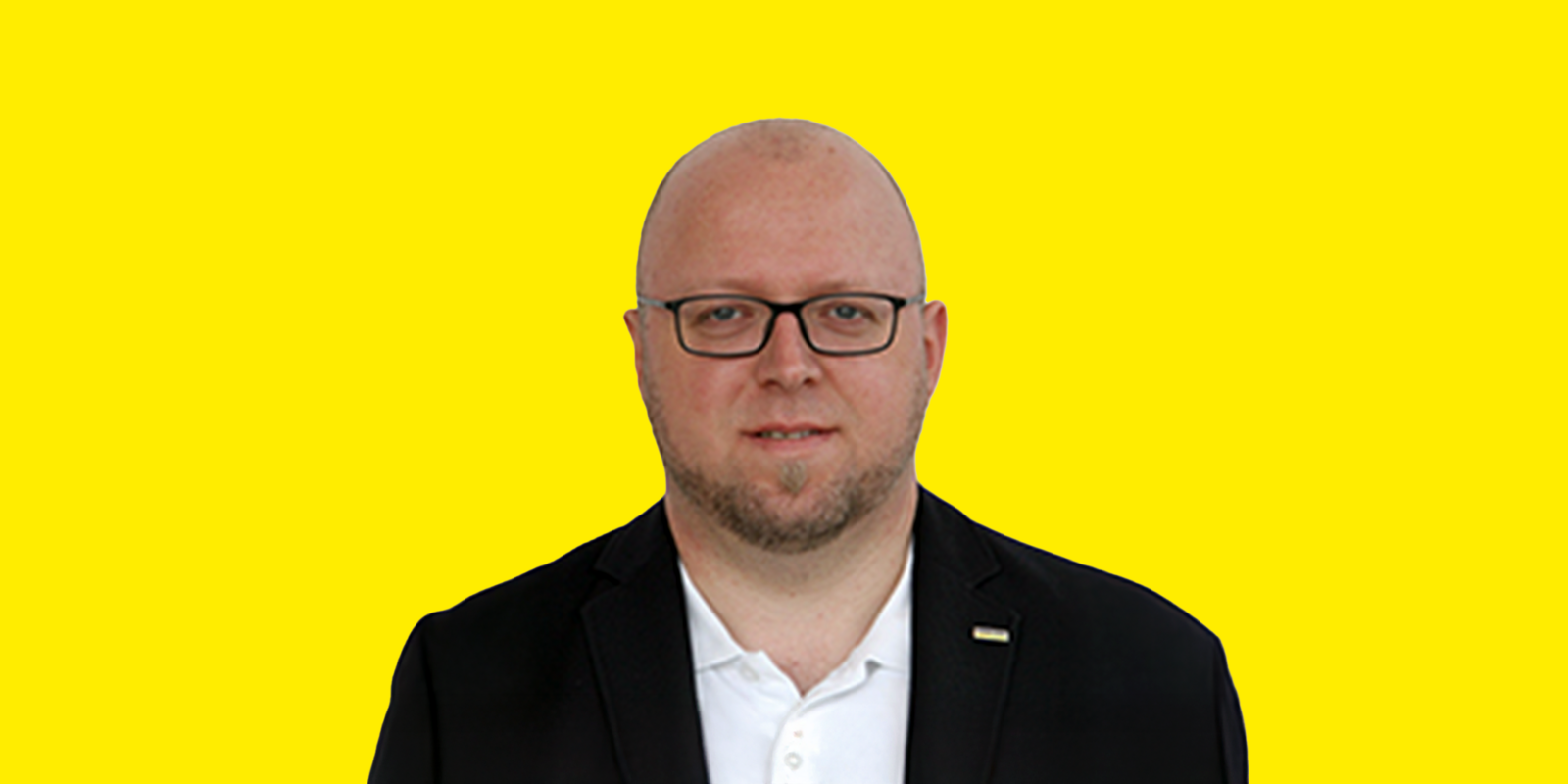 Christian Kaufmann Das Bild zeigt ein Porträt von Christian Kaufmann mit Brille, der direkt in die Kamera blickt. Er hat eine Glatze und einen kurzen Bart. Er trägt ein weißes Poloshirt und eine dunkle Jacke oder einen Blazer. Der Hintergrund ist ein schlichtes gelb.