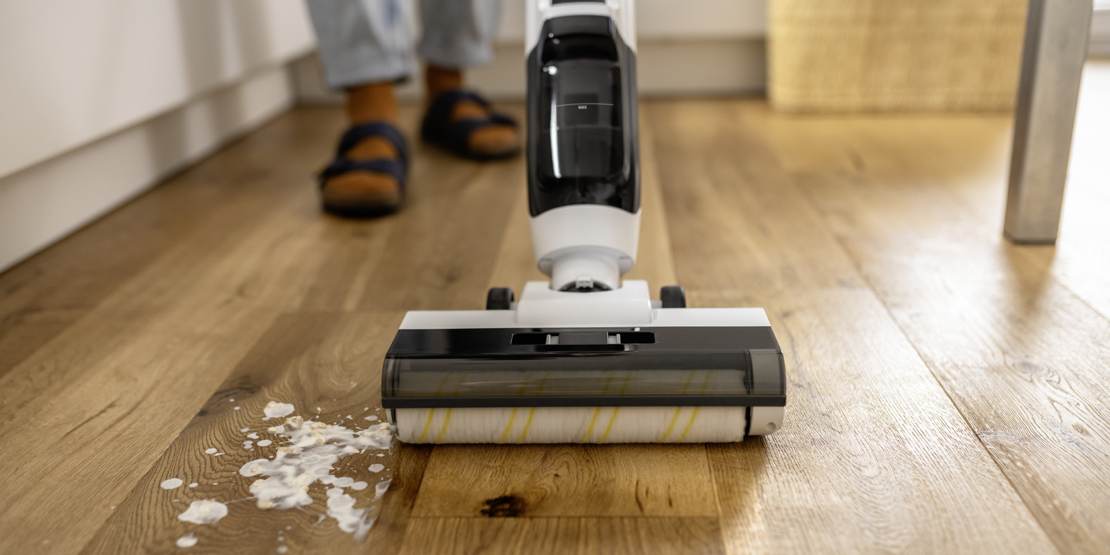 Saugwischer FCV 4 Ein moderner, weiß-schwarzer Bodenreinigungsgerät reinigt einen Holzboden. Links ist eine verschüttete, milchige Flüssigkeit zu sehen. Der Bereich rechts vom Gerät ist bereits sauber und glänzend. Im Hintergrund sind die Füße einer Person in Sandalen erkennbar.