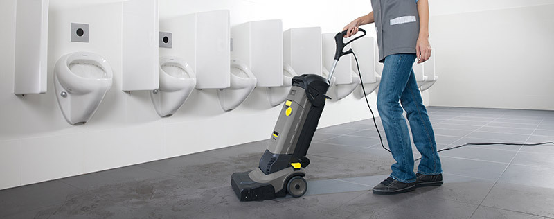 A person operates a walk-behind floor scrubber in a restroom