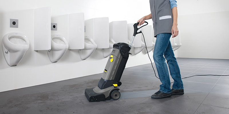 A worker cleans restroom floors with a compact walk-behind floor scrubber