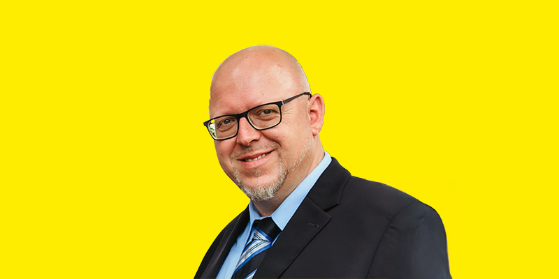 Christian Kaufmann Das Bild zeigt ein Porträt von Christian Kaufmann mit Brille, der direkt in die Kamera blickt. Er hat eine Glatze und einen kurzen Bart. Er trägt ein weißes Poloshirt und eine dunkle Jacke oder einen Blazer. Der Hintergrund ist ein schlichtes gelb.