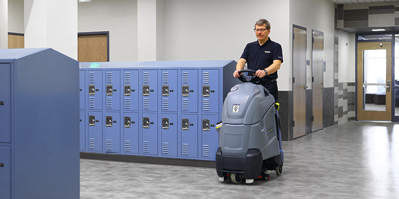 A worker cleans school floors with a Chariot stand-on floor scrubber