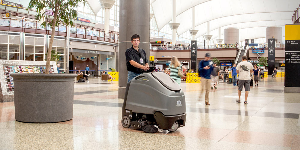A worker cleans floors at an airport using a Karcher Chariot stand-on floor scrubber