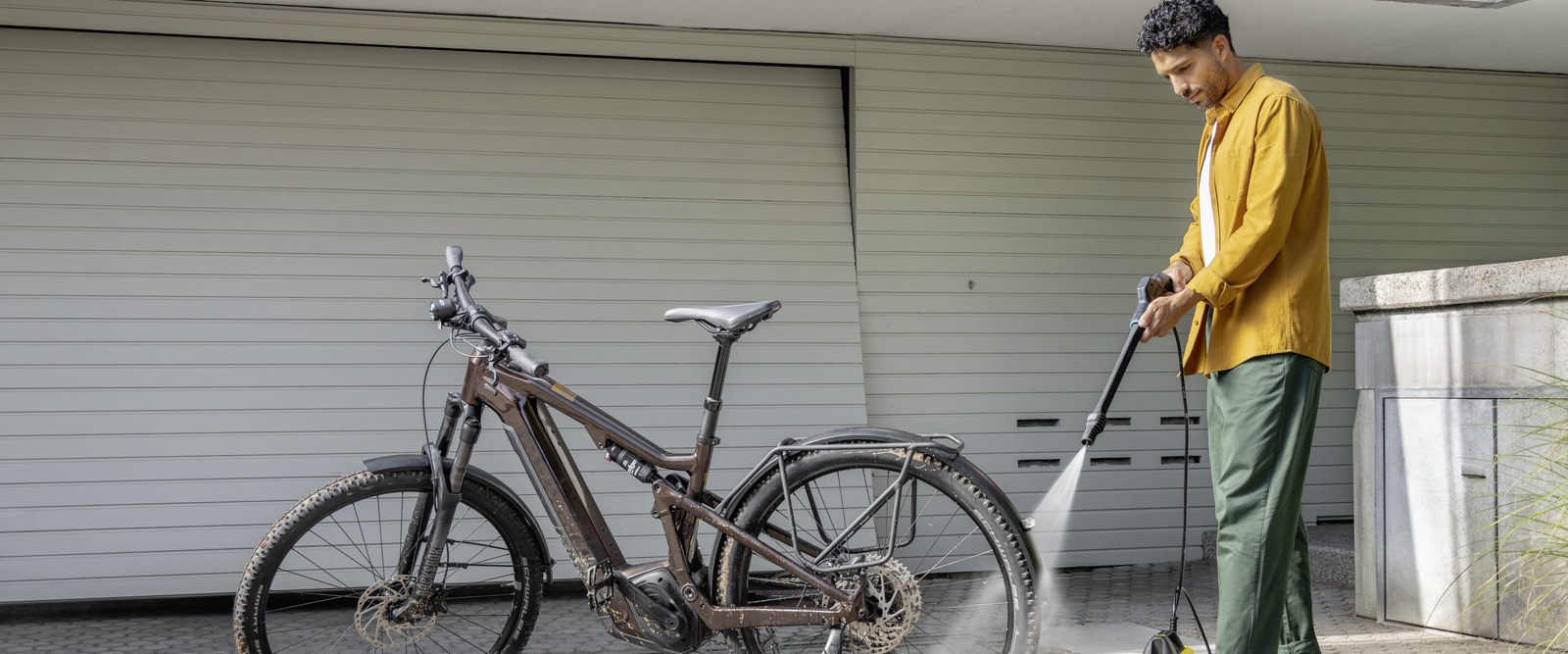 A man cleans his bicycle in front of the garage with a Kärcher Mobile Outdoor Cleaner.