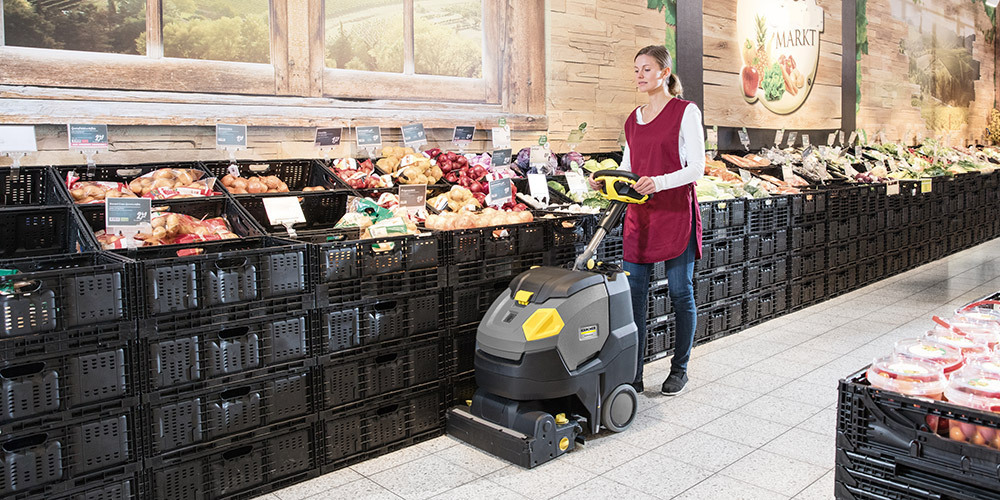 A woman cleaning at a grocery store with a walk-behind floor scrubber