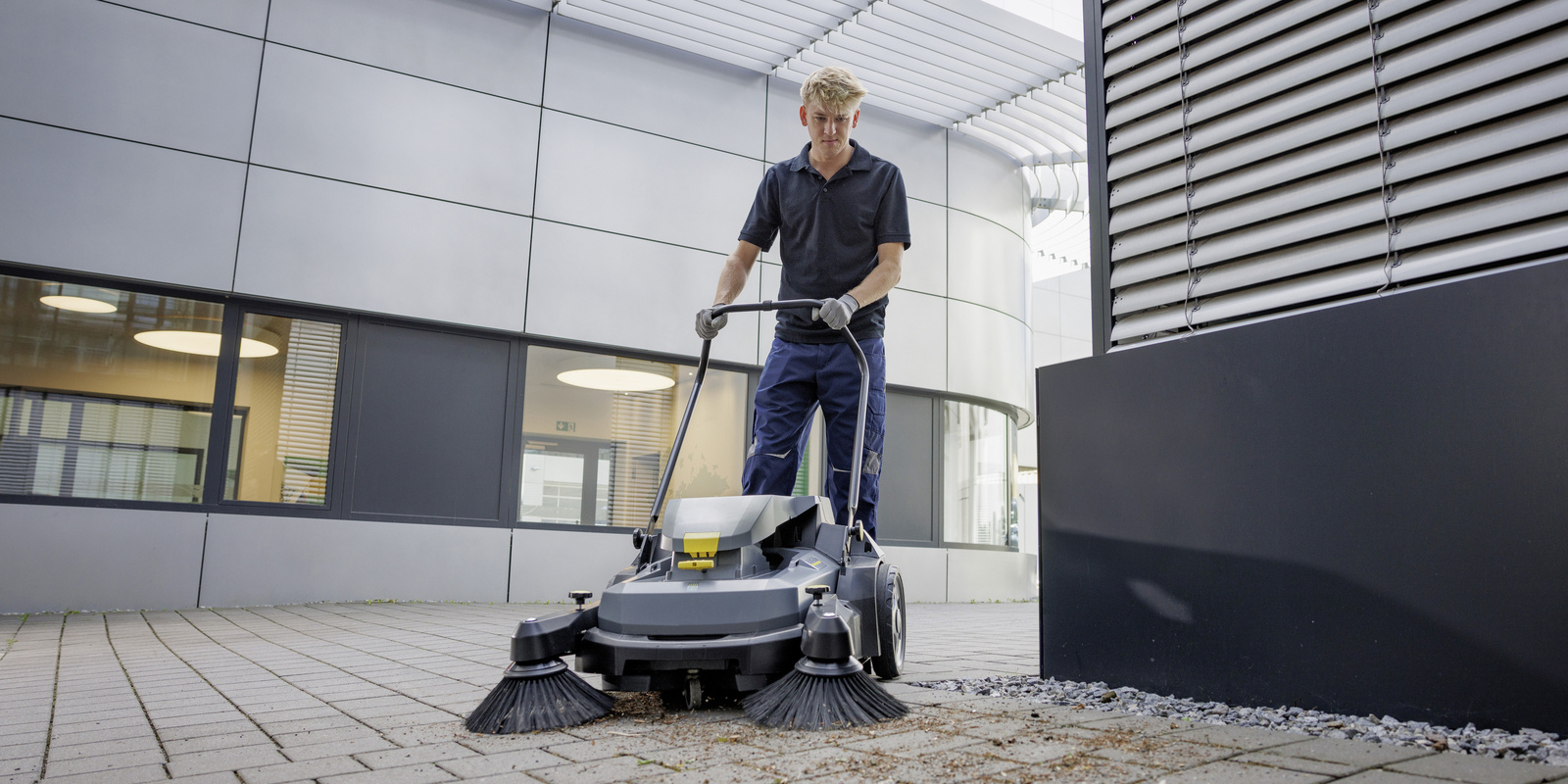 A worker sweeps outdoor concrete areas with a walk-behind floor sweeper