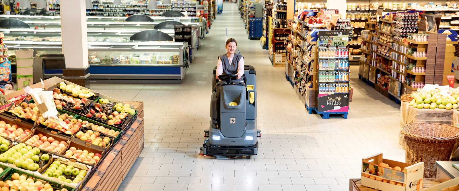 A person cleans grocery store tiled floors using a ride-on floor scrubber