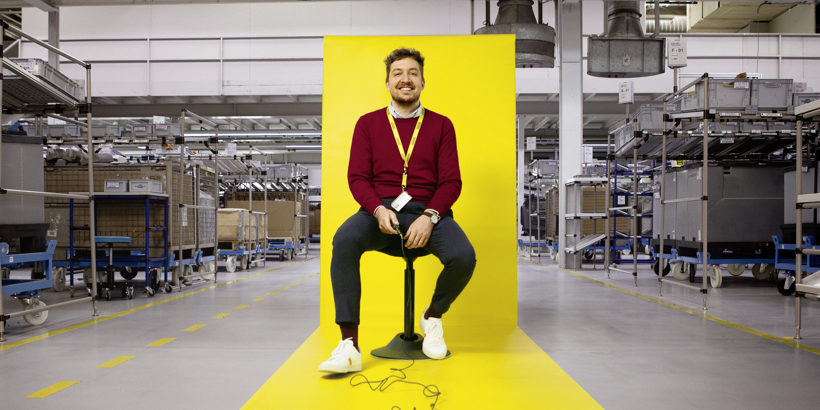 A worker sits on a stool with a yellow backdrop within a factory 
