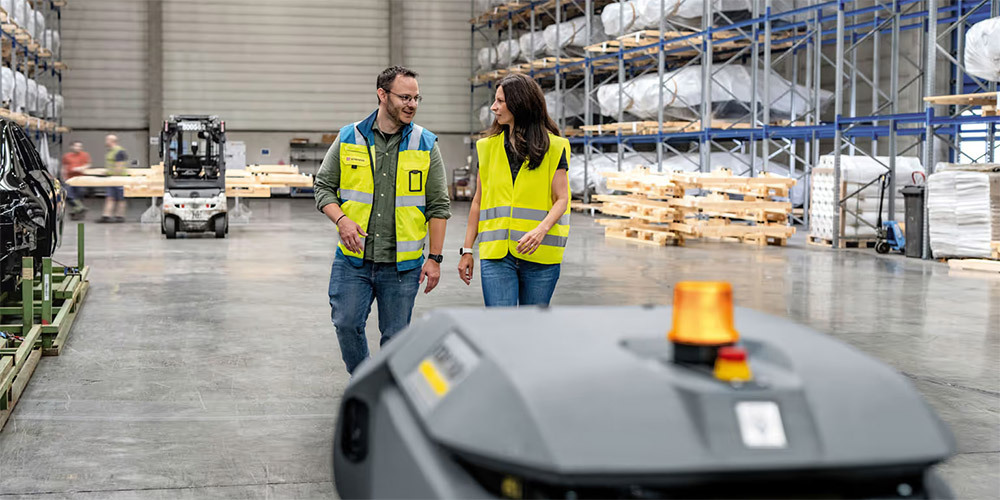 Workers walk in a warehouse near a working KIRA B 50 autonomous floor scrubber.