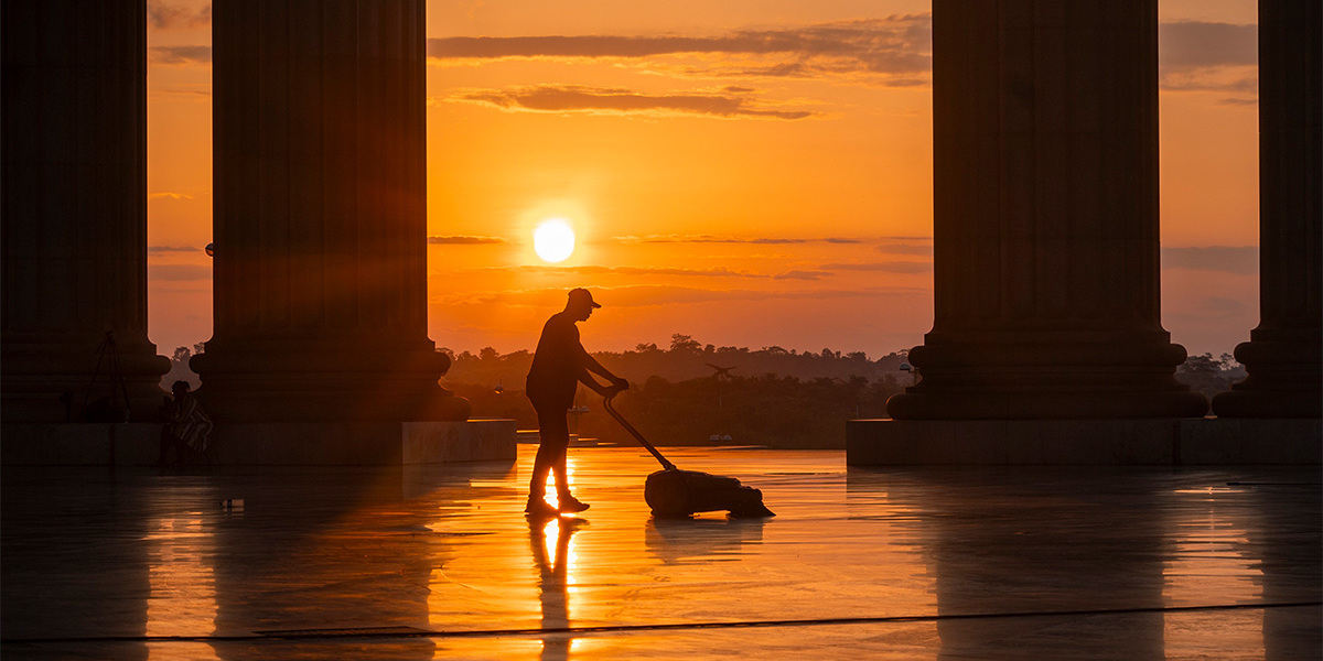 Silhouette of a person using a Kärcher cleaning machine to clean a large stone plaza between columns at sunrise