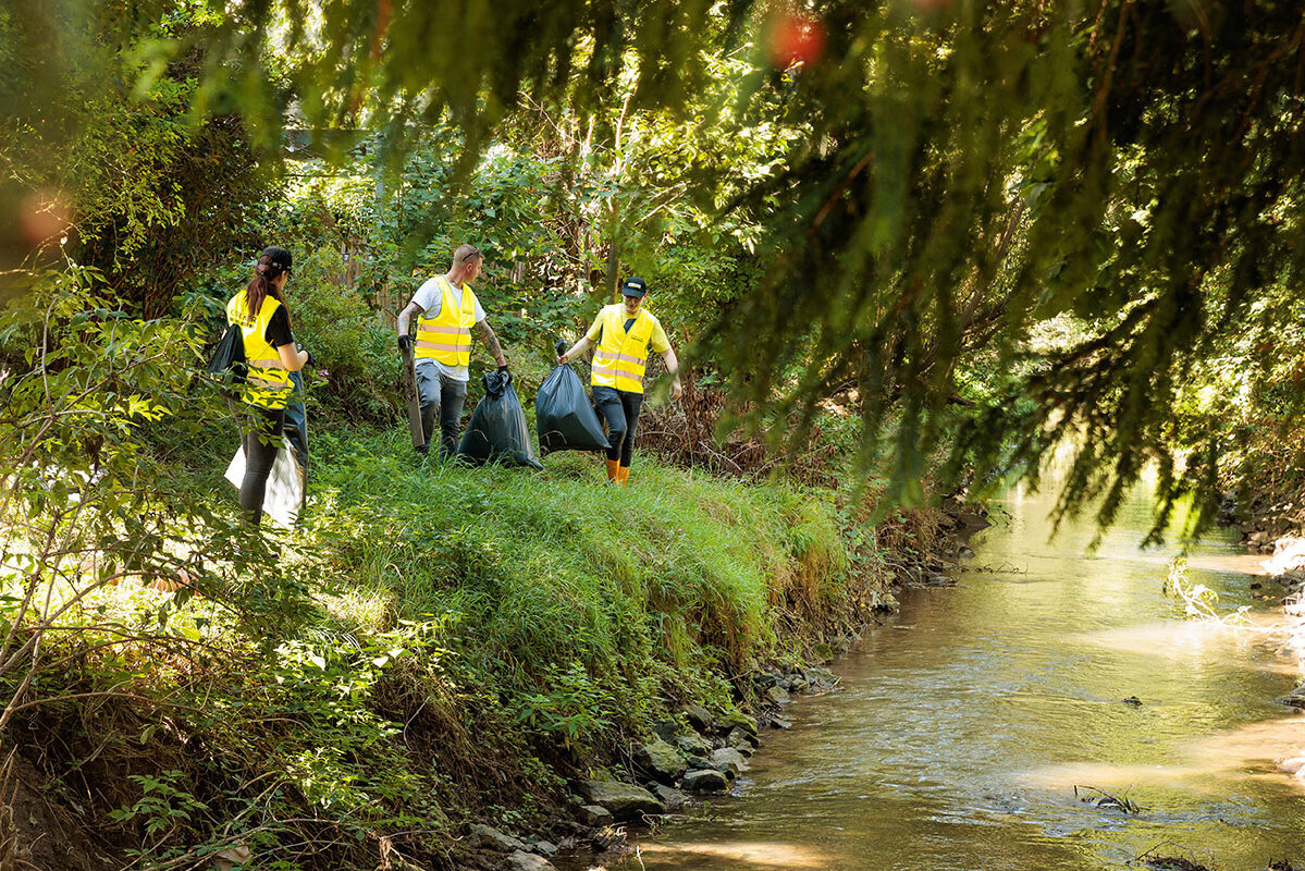 Three volunteers in yellow safety vests collecting trash bags along a green, overgrown riverbank as part of a Kärcher clean-up initiative