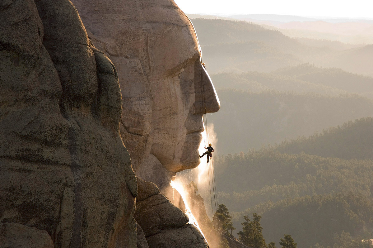 A worker rappelling down the George Washington face at Mount Rushmore during a high-pressure cleaning operation, with forested hills in the background