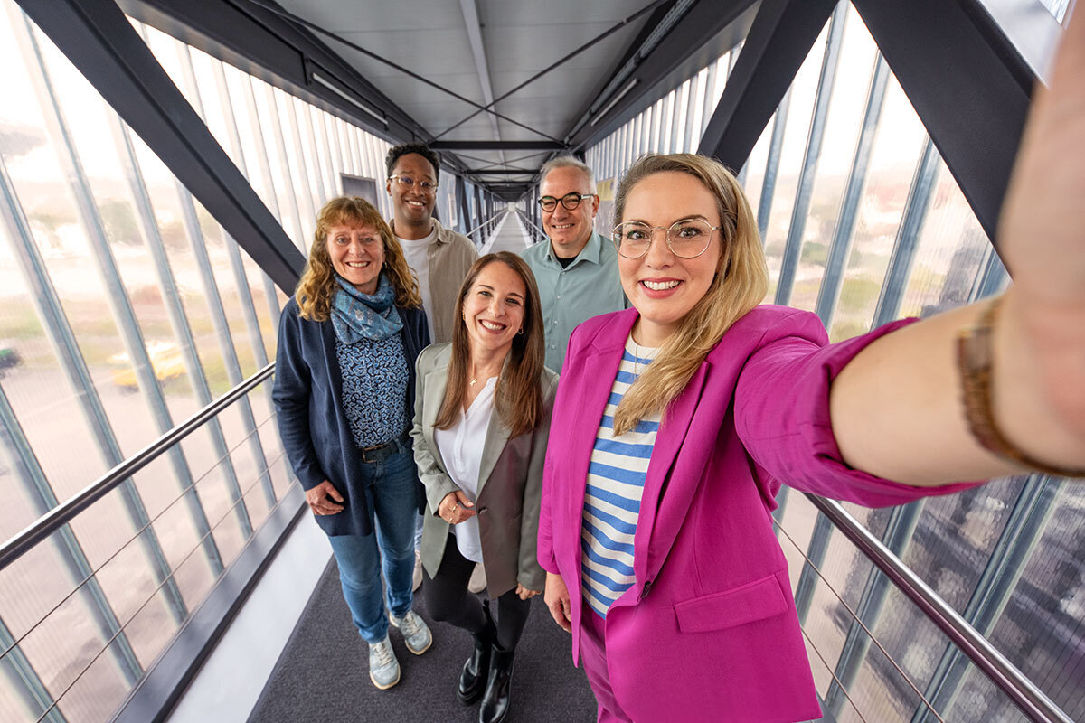 Five smiling Kärcher colleagues posing for a group selfie on a modern glass-and-steel office walkway