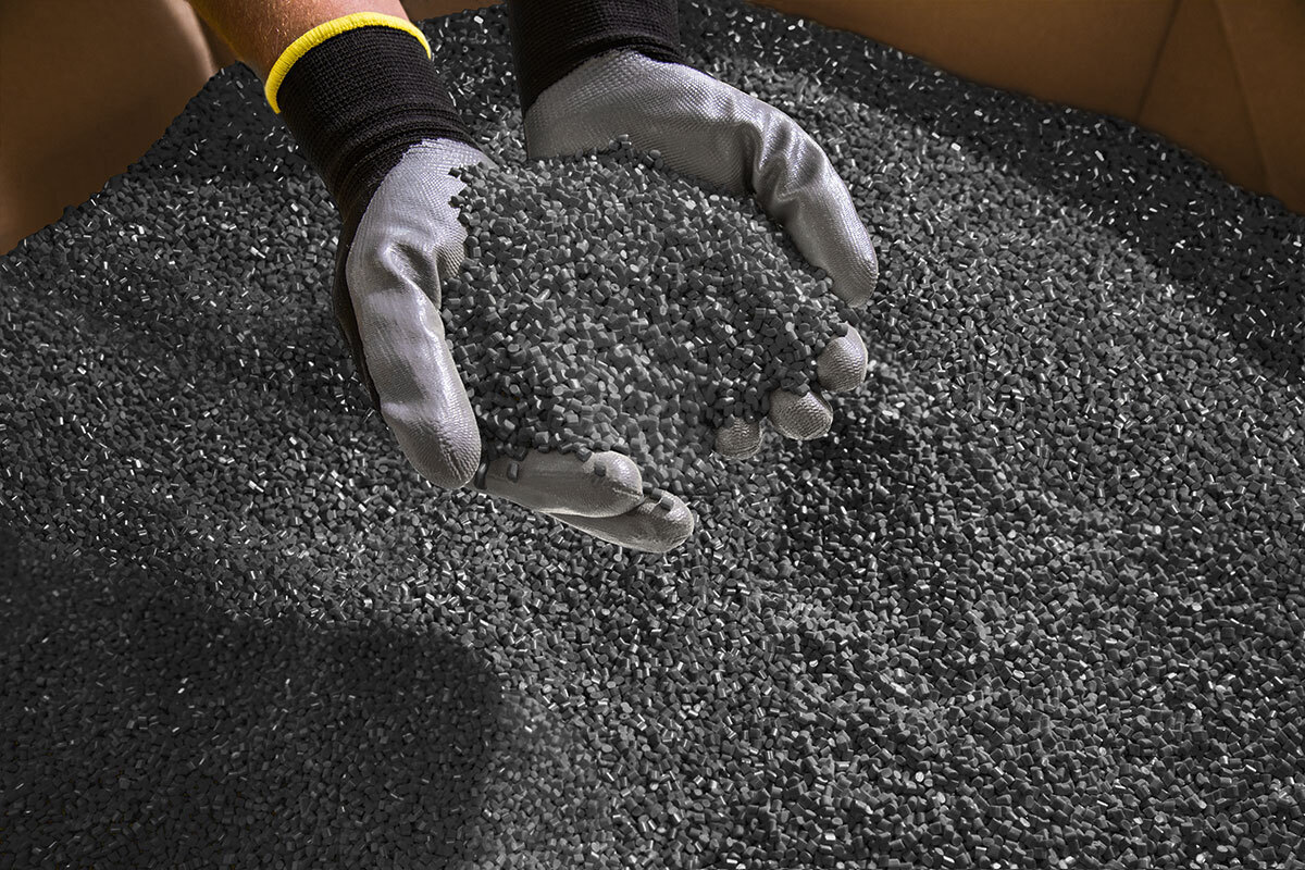 Gloved hands holding a scoop of dark recycled plastic granulate, with a large surface of the material spread out underneath