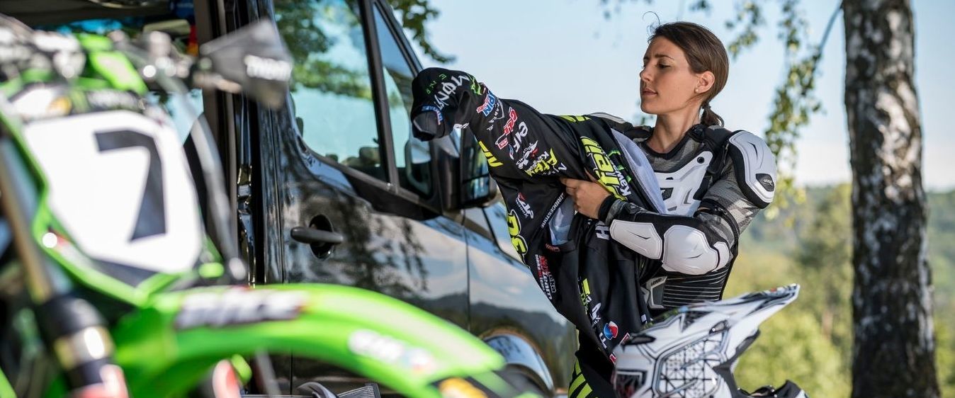 Motocrossfahrerin Vanessa Helpert zieht ihr Trikot über die Schutzausrüstung. Auf dem Trikot sind die Sponsorenlogos zu sehen.